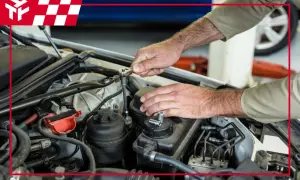 Mechanik naprawia silnik samochodu, używając klucza nasadowego. W tle widać logo z czerwoną kratką, przypominające flagę wyścigową, sugerujące serwis samochodowy w **nagar**.