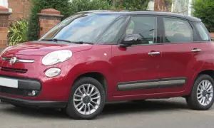 Czerwony Fiat 500 L z czarnym dachem zaparkowany przy ceglanym murze i zieleni.