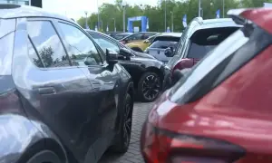 Rzędy samochodów, w tym czerwony i szary SUV, czekają na swoich właścicieli. W tle widać bramę z logo, sugerującą salon samochodowy lub wydarzenie motoryzacyjne.