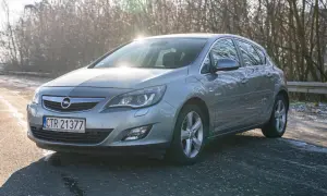 Szary Opel Astra J 1.4 Turbo na tle drzew, z widocznym słońcem odbijającym się od karoserii.
