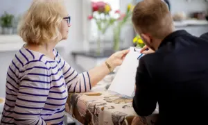 Kobieta z blond włosami i okularami podaje dokument młodemu mężczyźnie. Prawdopodobnie wypełniają wniosek o rejestrację pojazdu pdf.