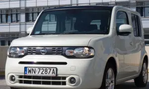 Biały Nissan Cube z polskimi tablicami rejestracyjnymi WN 7287A zaparkowany przed nowoczesnym budynkiem.