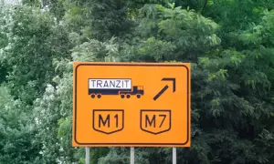 Pomarańczowy znak drogowy w drodze na Węgry. Informuje o tranzycie ciężarówek na autostrady M1 i M7.