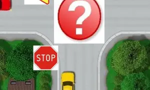 Czerwony samochód zbliża się do skrzyżowania, gdzie znak a7 sugeruje ustąpienie pierwszeństwa. Na drodze znak STOP.