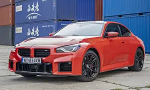 Pomarańczowe BMW M2 z czarnymi felgami stoi na tle niebieskich kontenerów. Sprawdź aktualną bmw m2 cenę!