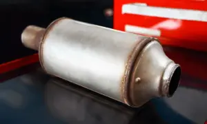 Metalowy tłumik, część układu wydechowego, leży na ciemnej powierzchni. W tle widać czerwony element, być może skrzynkę narzędziową. To element systemu GPF.