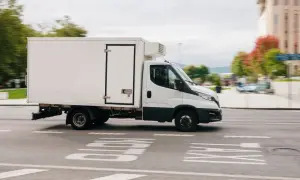 Biała chłodnia na podwoziu Iveco Daily, idealna do transportu wymagającego odpowiedniego prawa jazdy C1, jedzie po miejskiej ulicy.