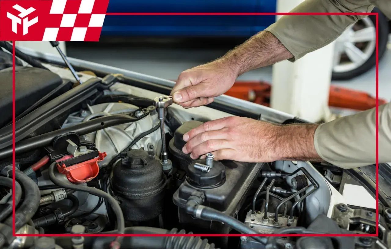 Mechanik naprawia silnik samochodu, używając klucza nasadowego. W tle widać logo z czerwoną kratką, przypominające flagę wyścigową, sugerujące serwis samochodowy w **nagar**.