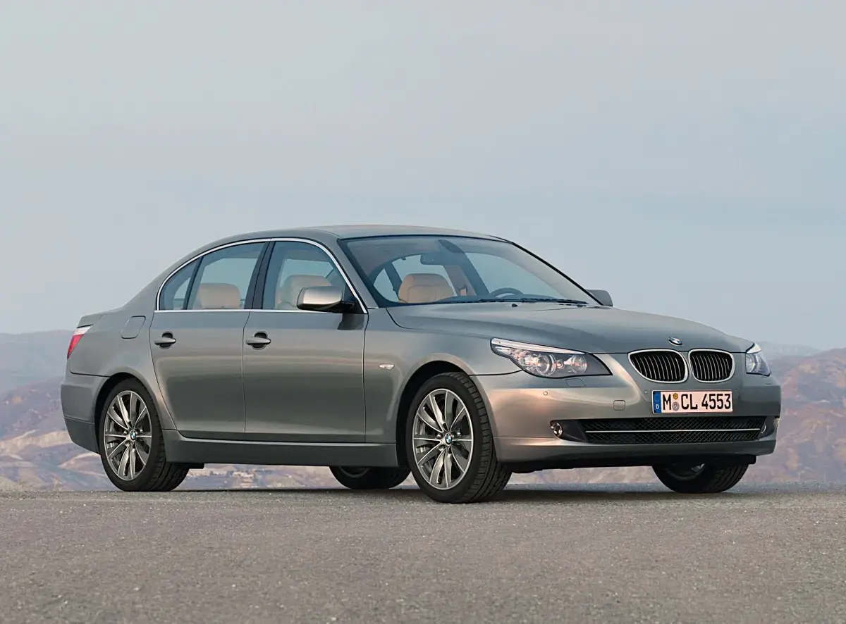 Szary sedan BMW 5 E60 z jasnym wnętrzem stoi na pustej drodze, z górami w tle.