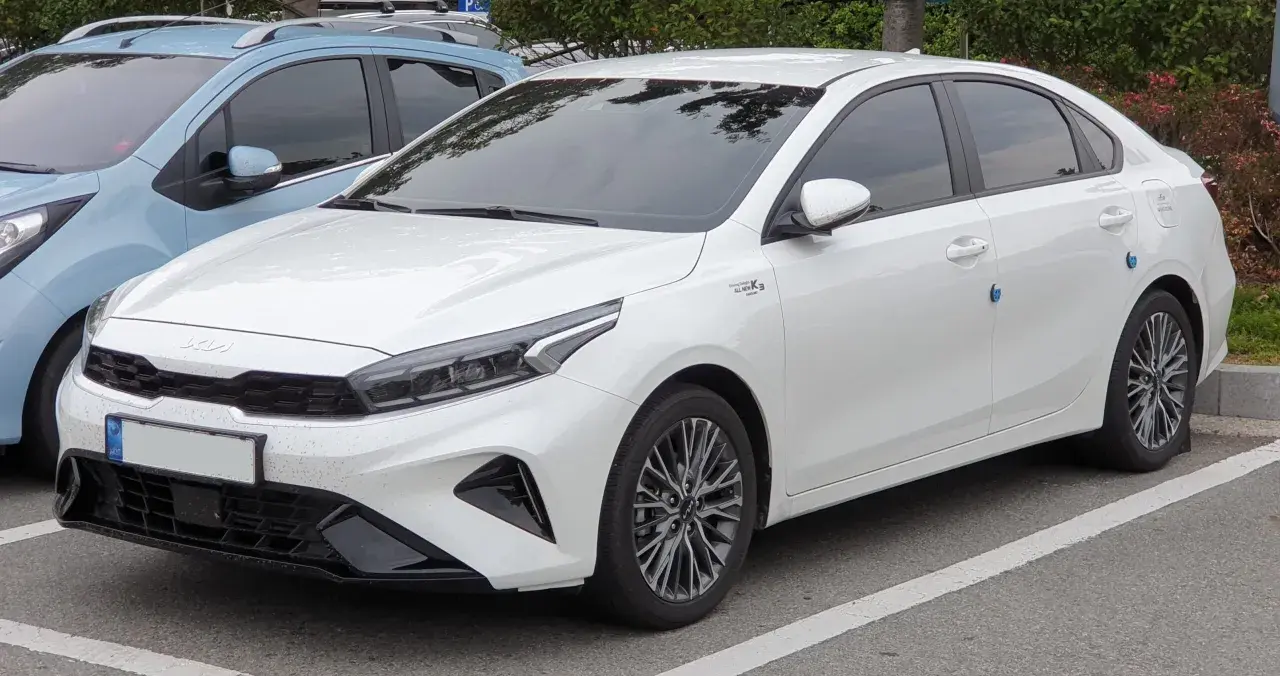 Biały sedan Kia Forte zaparkowany na parkingu, widok z przodu i z boku.