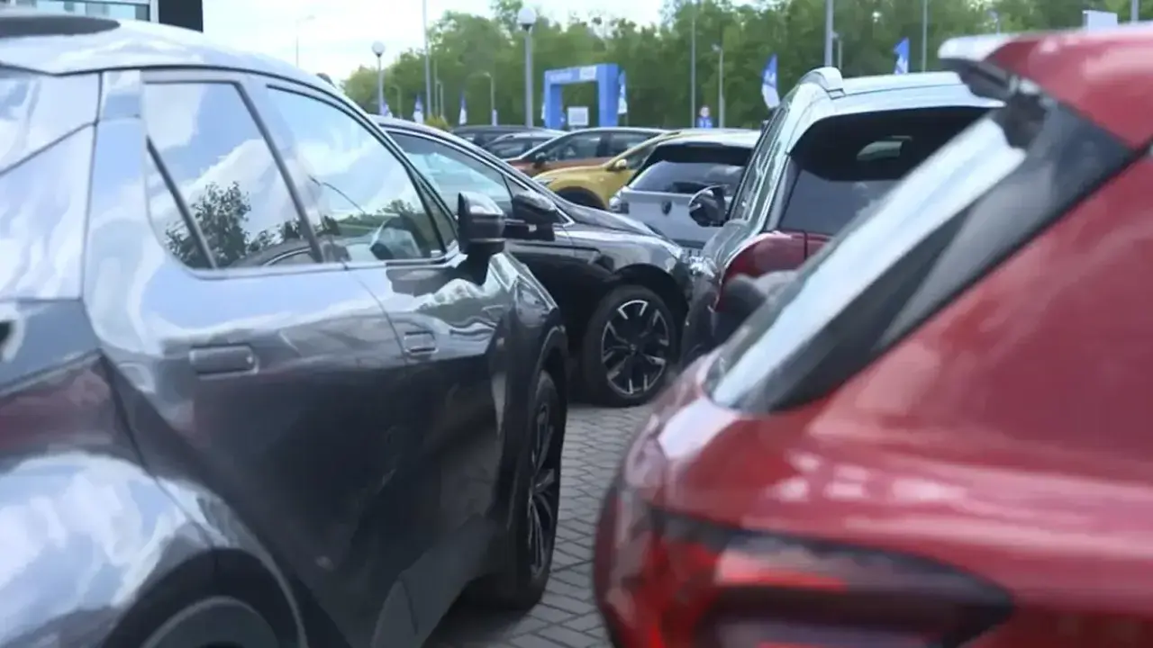 Rzędy samochodów, w tym czerwony i szary SUV, czekają na swoich właścicieli. W tle widać bramę z logo, sugerującą salon samochodowy lub wydarzenie motoryzacyjne.