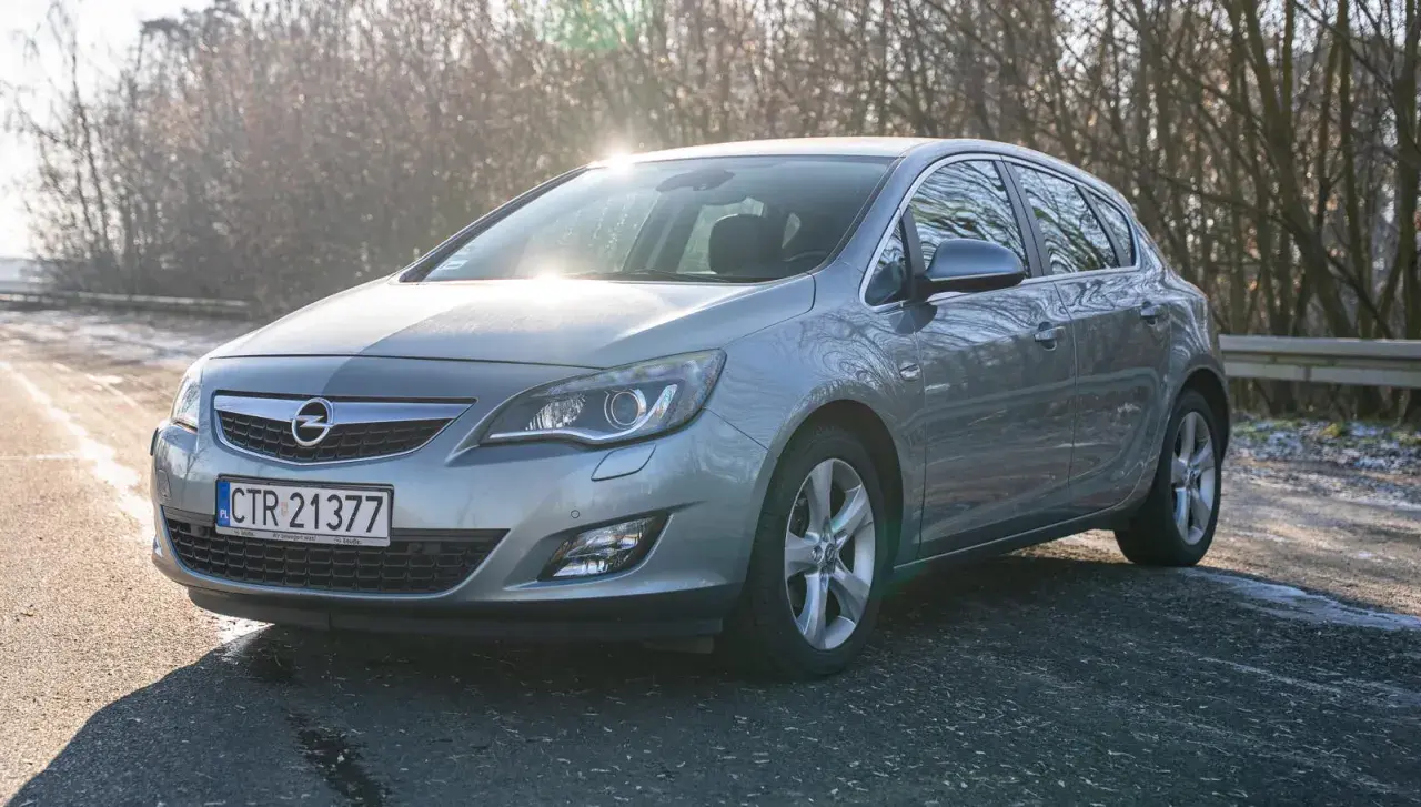Szary Opel Astra J 1.4 Turbo na tle drzew, z widocznym słońcem odbijającym się od karoserii.