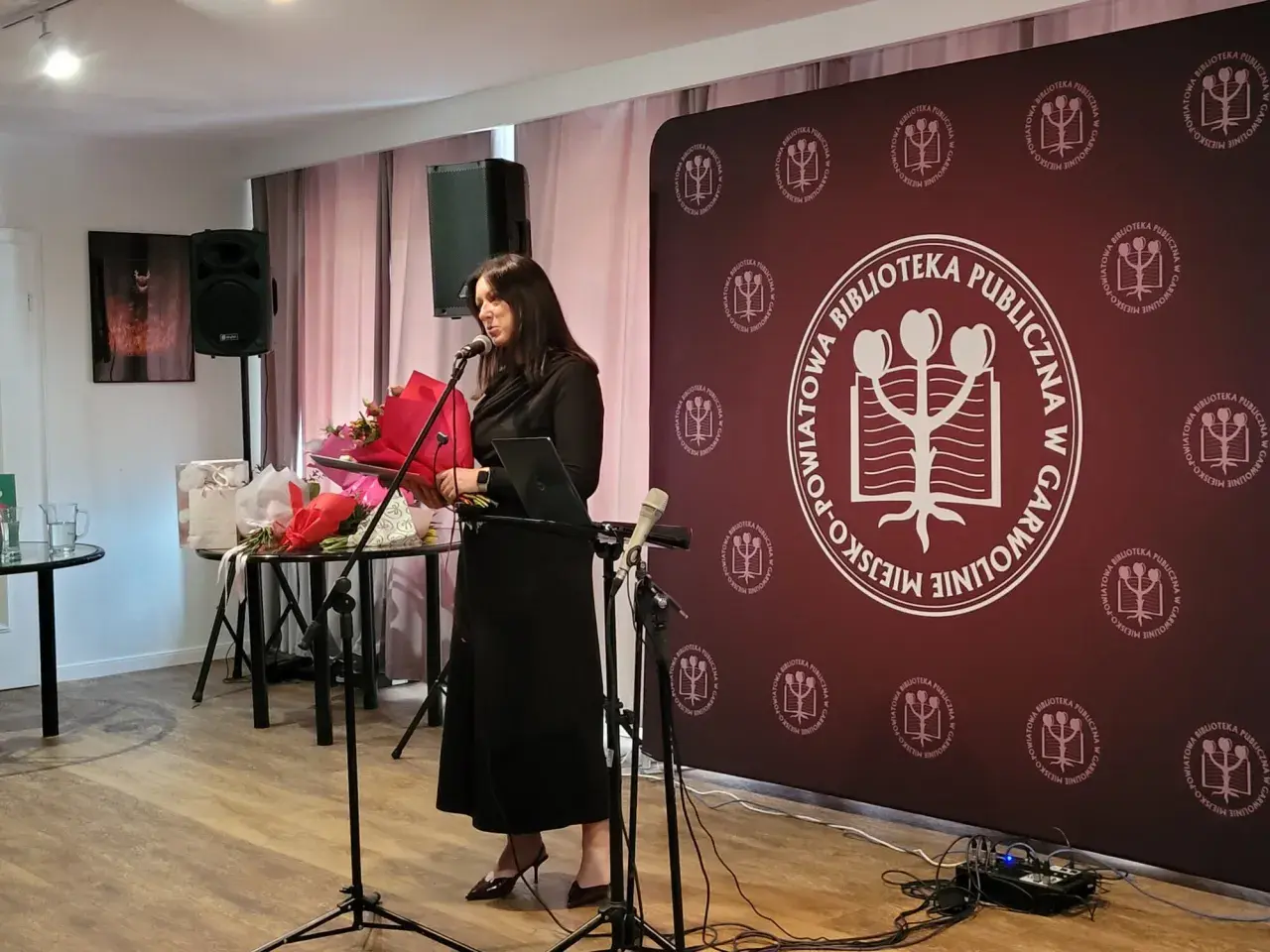 Kobieta przemawia na tle baneru z logo biblioteki, podczas wydarzenia w Garwolinie. Wgm rejestracja.