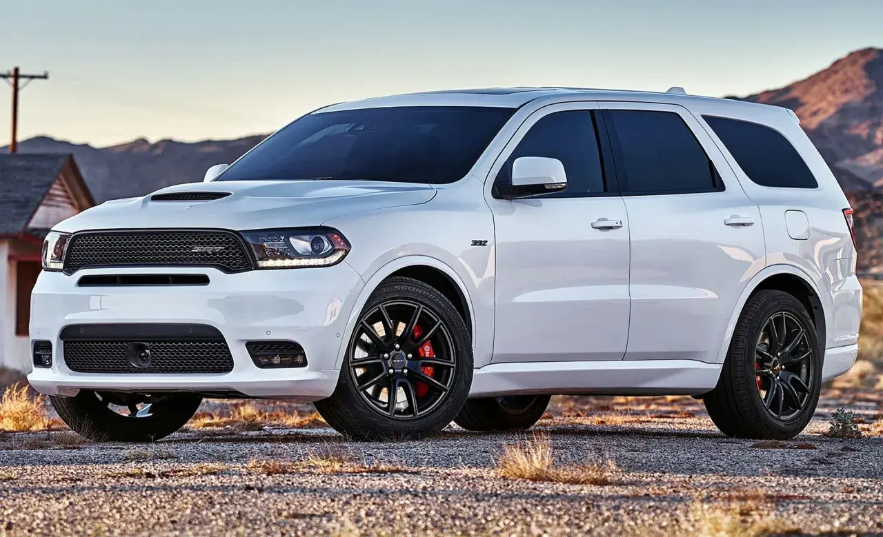Biały Dodge Durango 3.6 SRT z czarnymi felgami i czerwonymi zaciskami hamulcowymi stoi na żwirowej drodze, w tle góry i opuszczony dom.