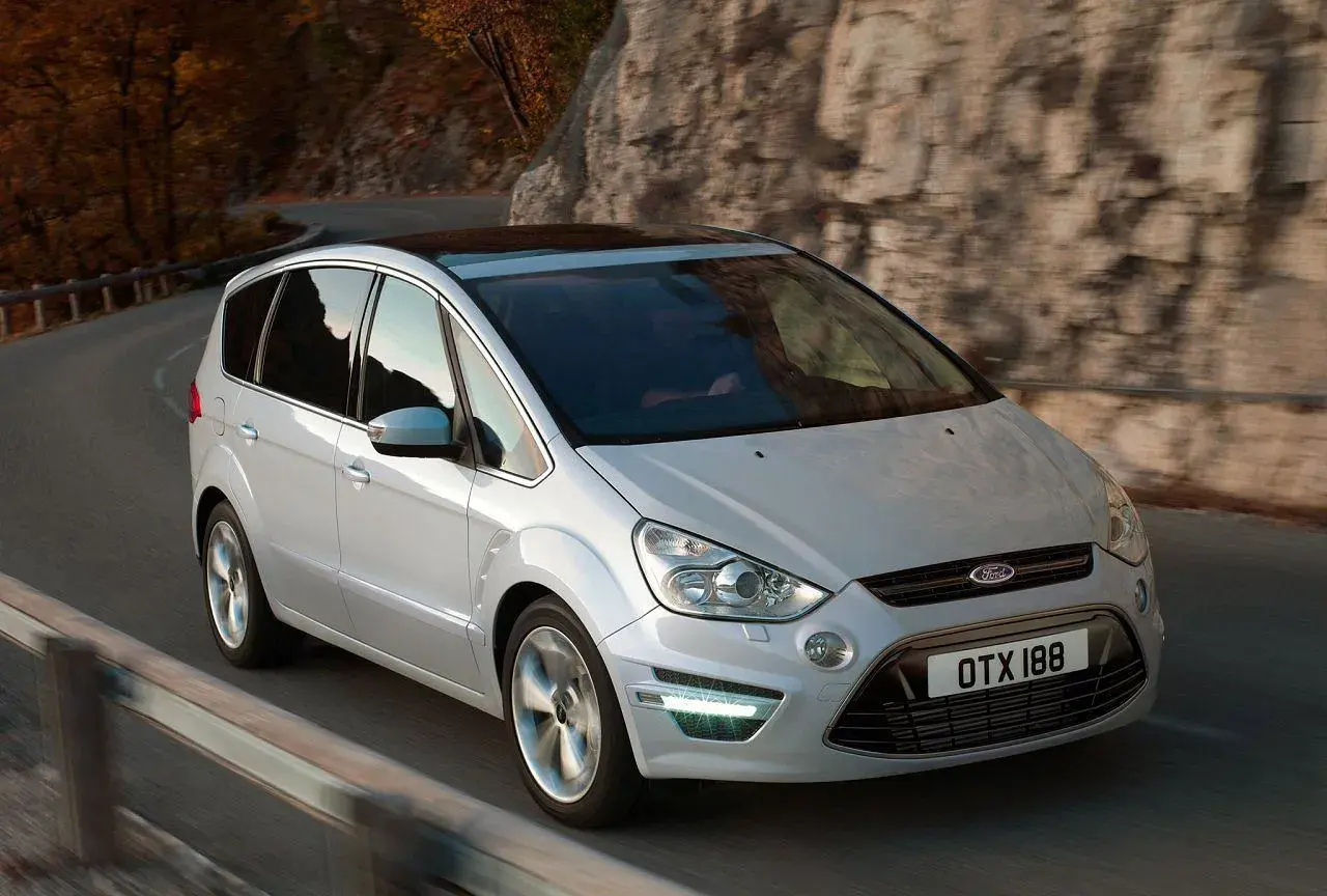 Biały Ford S-Max 2.0 TDCI na krętej drodze wśród jesiennych drzew i skał.