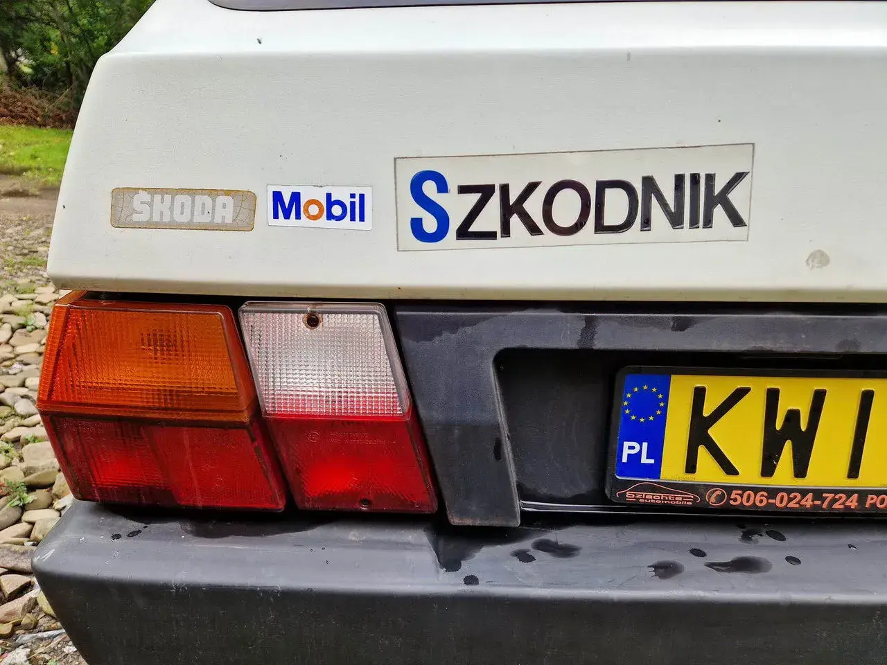 Tył białego samochodu Skoda z naklejkami "Mobil" i "Szkodnik". Widoczna tablica rejestracyjna KWI.