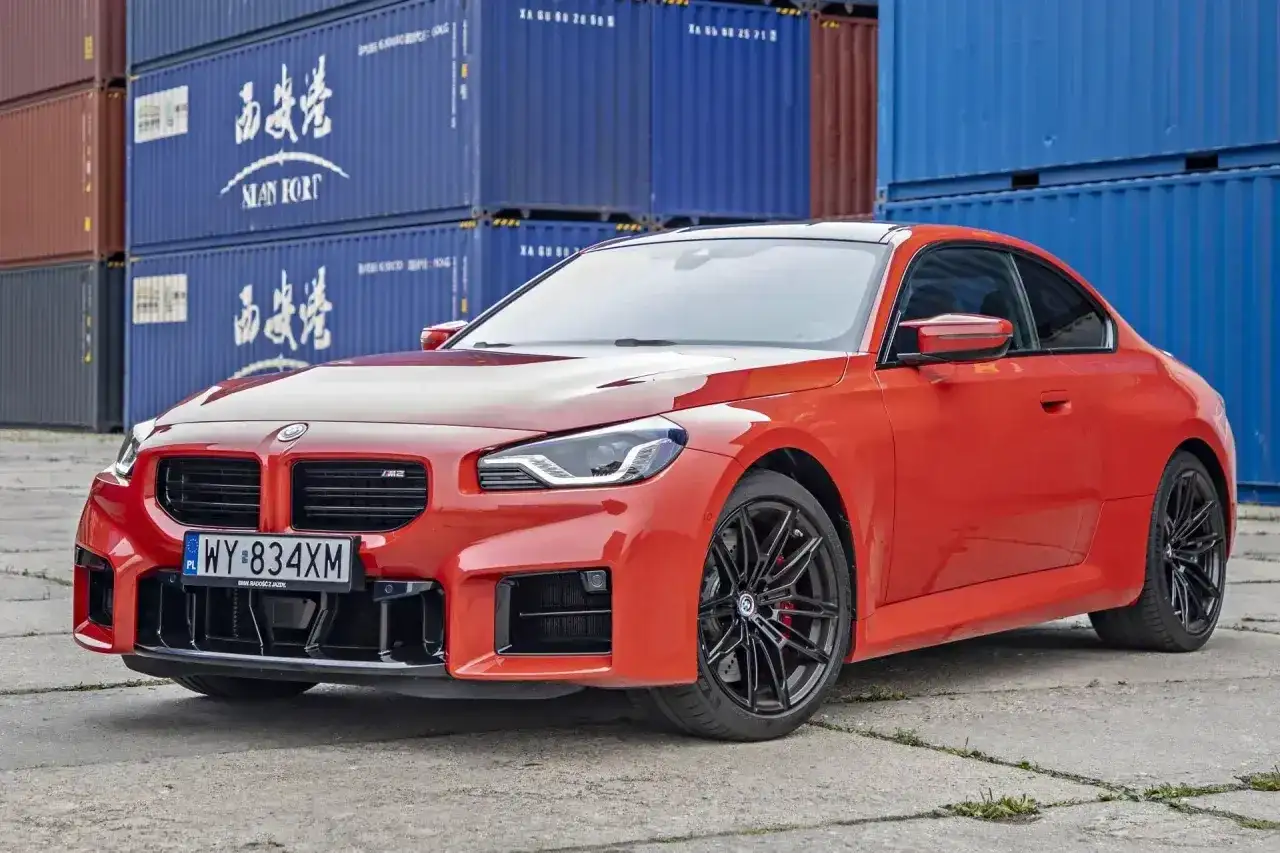 Pomarańczowe BMW M2 z czarnymi felgami stoi na tle niebieskich kontenerów. Sprawdź aktualną bmw m2 cenę!
