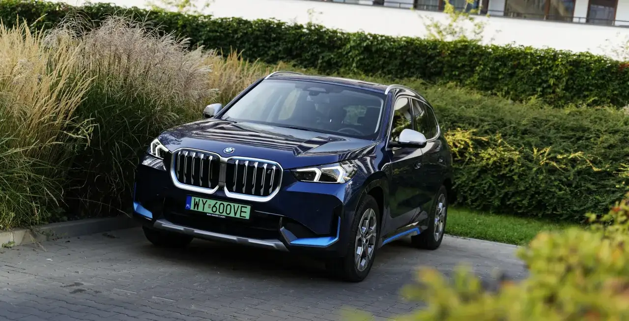 Ciemnoniebieskie BMW X1 na podjeździe, otoczone zielenią. Sprawdź aktualną bmw x1 cena i ofertę.
