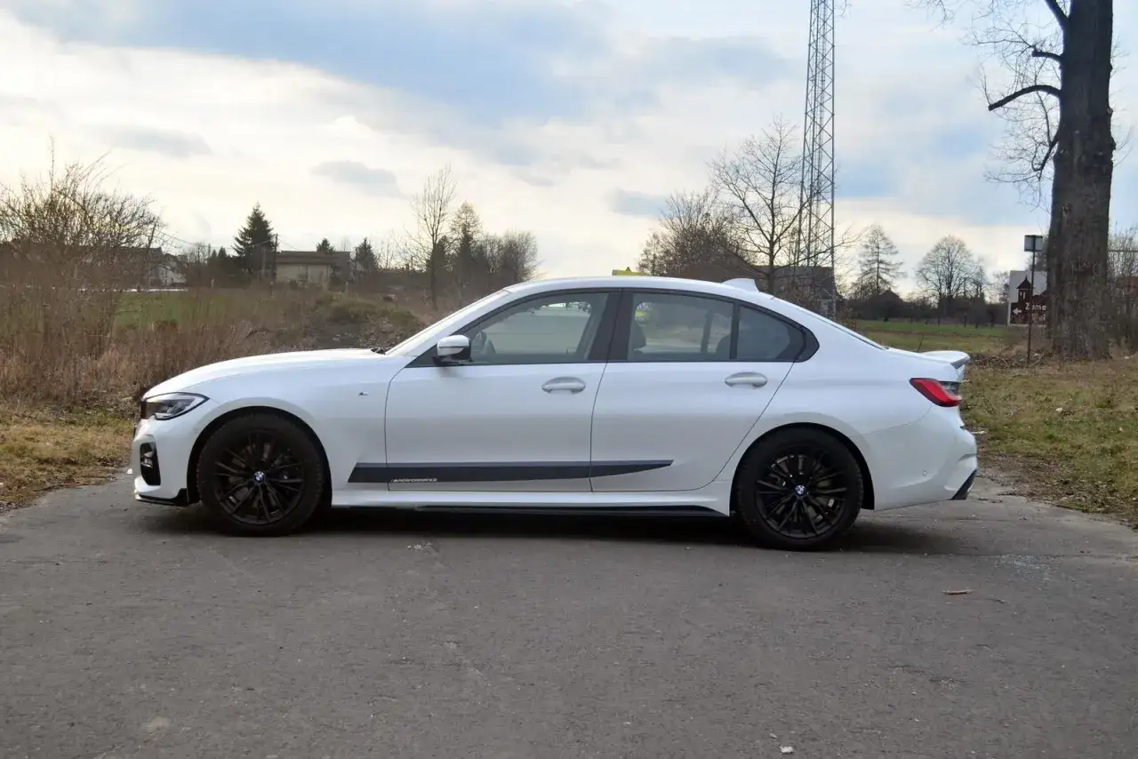 Białe BMW 318d z czarnymi felgami i spoilerem, zaparkowane na drodze.