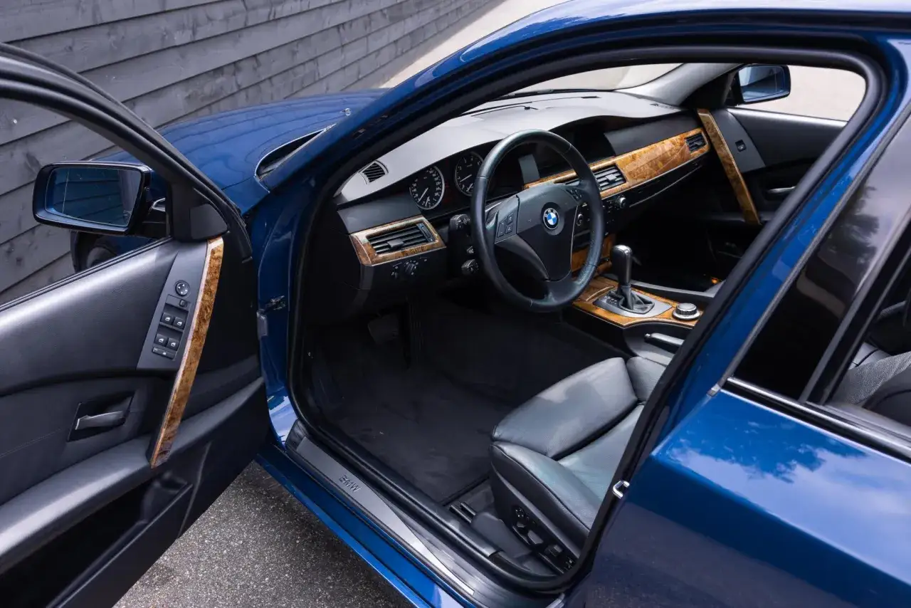 Wnętrze BMW 5 E60 z drewnianymi wstawkami i czarną skórzaną tapicerką. Widoczna kierownica, deska rozdzielcza i fotel kierowcy.