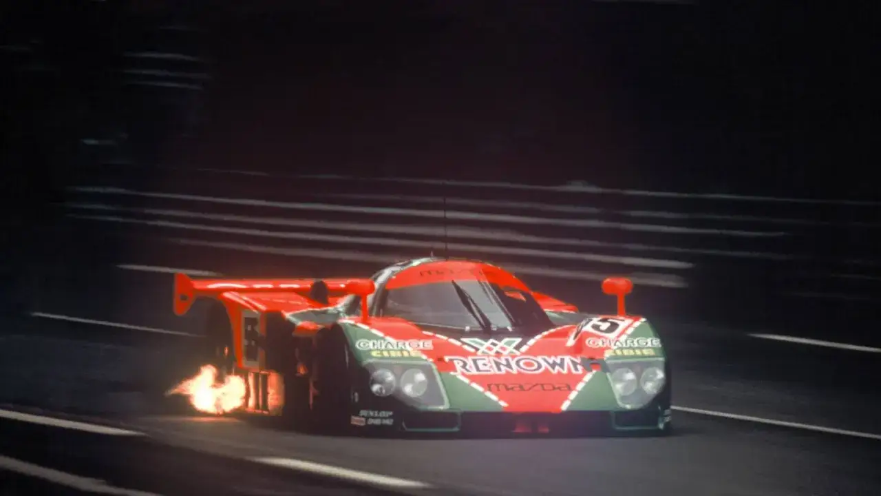 Wyścigowa Mazda 787B z płomieniami wydobywającymi się z tyłu, pędząca po torze nocą.
