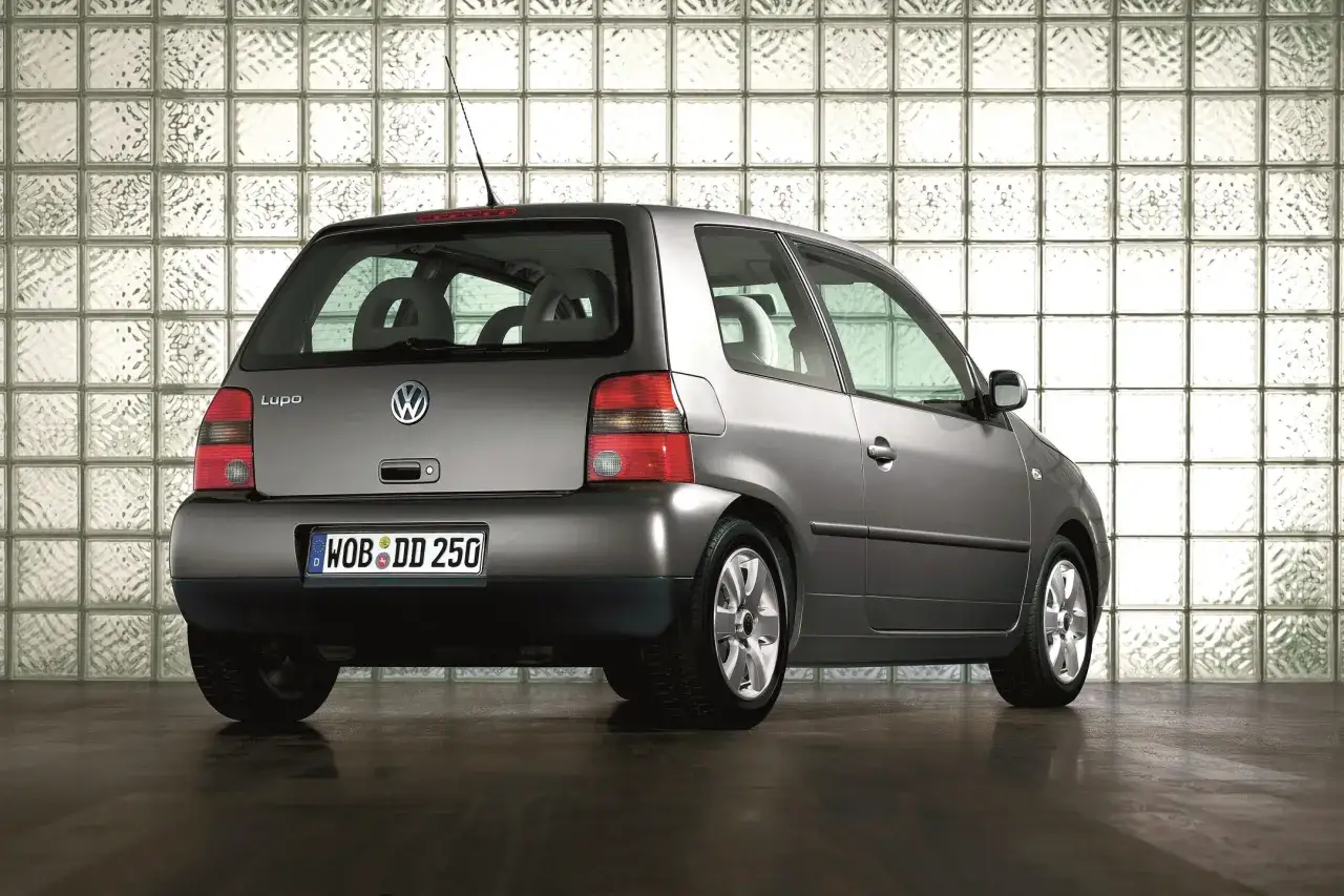Szary Volkswagen Lupo stoi na ciemnej podłodze przed ścianą z cegieł szklanych.