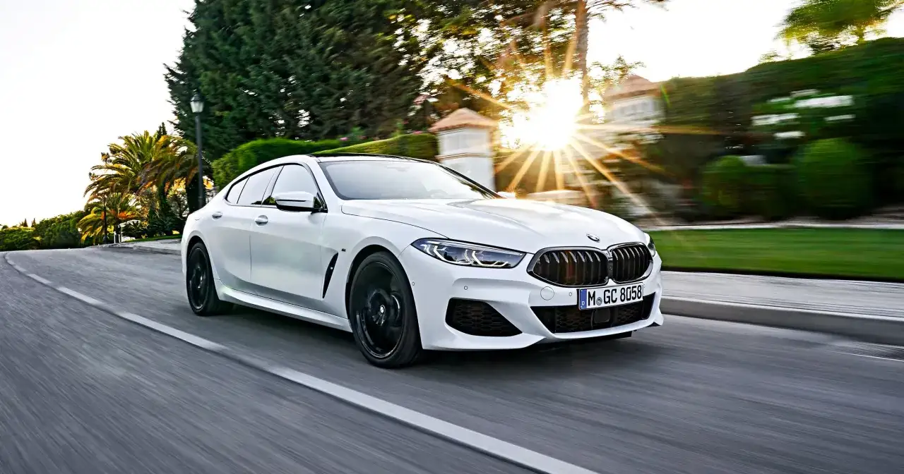 Białe BMW 840i sunie po drodze w promieniach zachodzącego słońca, otoczone bujną zielenią.