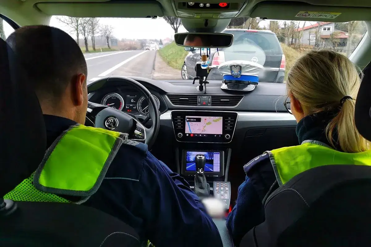 Policjanci w radiowozie, gotowi wystawić mandat za brak prawa jazdy. Na desce rozdzielczej czapka policyjna i nawigacja.