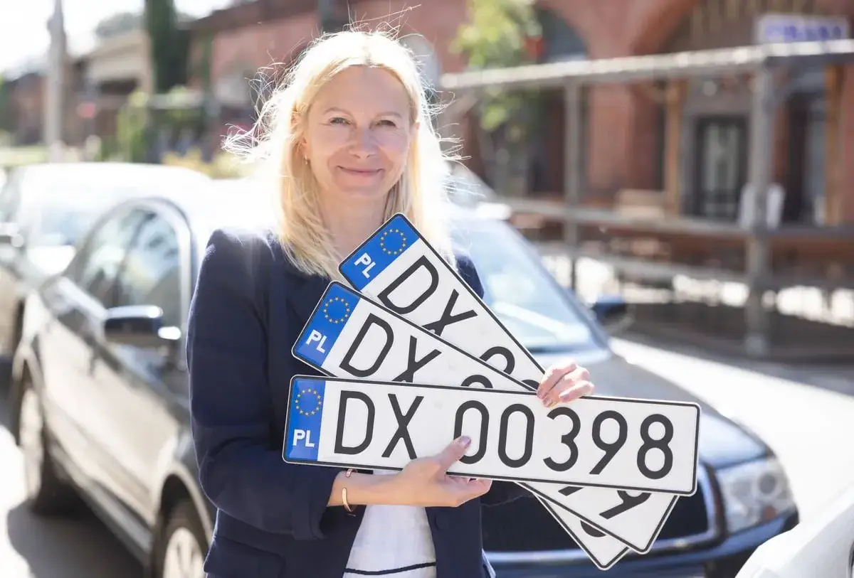 Kobieta trzyma tablice rejestracyjne z oznaczeniem DX. Nowe tablice ułatwiają dx rejestrację pojazdów.