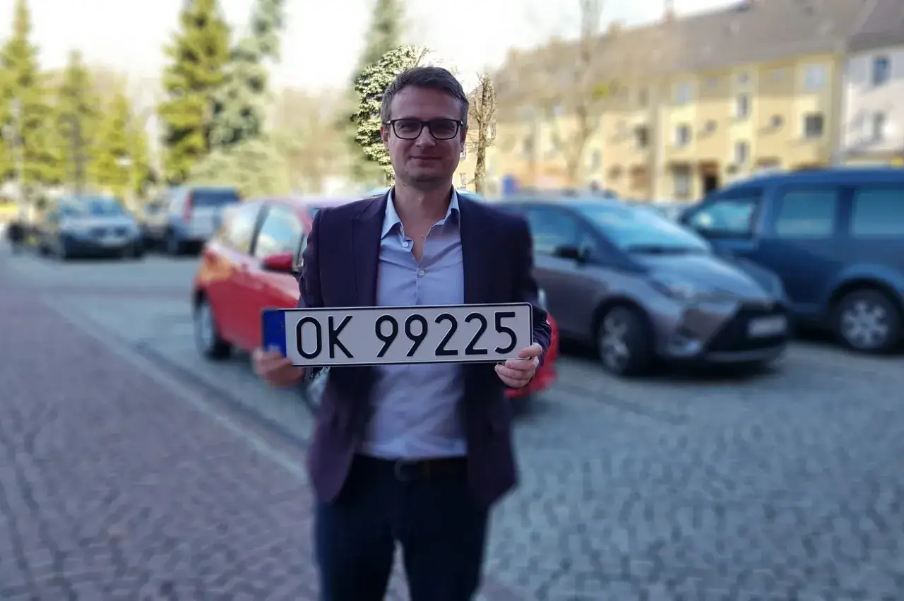 Mężczyzna w garniturze trzyma tablicę rejestracyjną OK 99225. Za nim widać zaparkowane samochody.