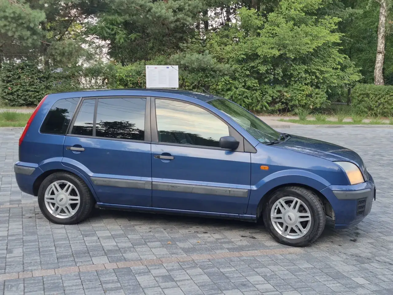 Niebieski Ford Fusion 1.4 benzyna zaparkowany na kostce brukowej, otoczony zielenią.