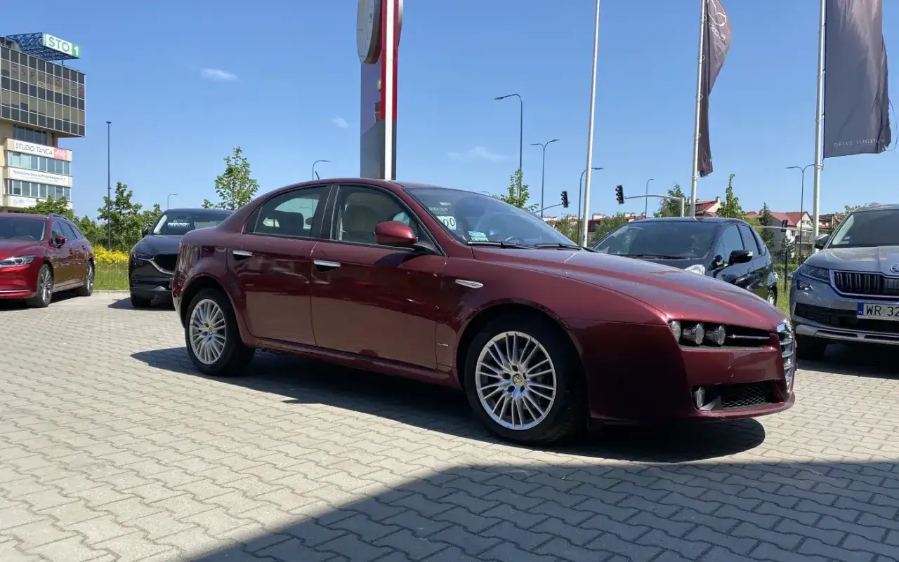 Bordowy Alfa Romeo 159 1.9 JTD na parkingu przed salonem samochodowym.