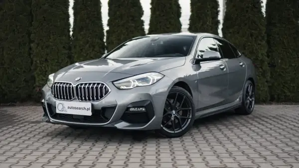 Szary BMW 218i z czarnymi felgami stoi przed żywopłotem.
