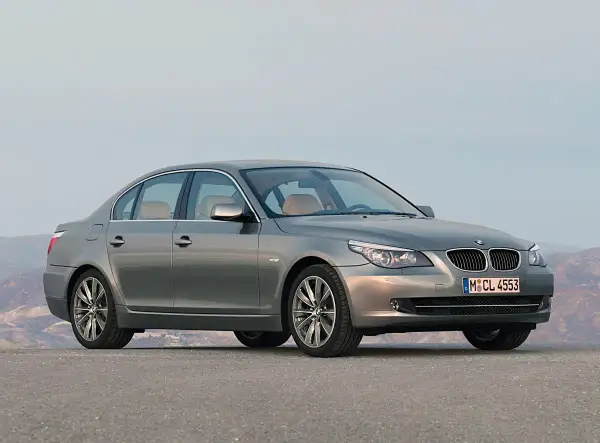 Szary sedan BMW 5 E60 z jasnym wnętrzem stoi na pustej drodze, z górami w tle.