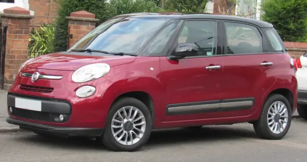 Czerwony Fiat 500 L z czarnym dachem zaparkowany przy ceglanym murze i zieleni.