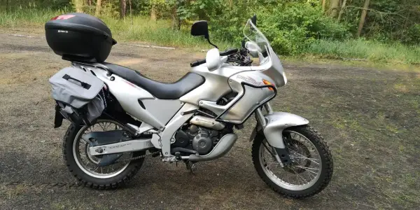Srebrny motocykl Aprilia Pegaso 650 z kufrem i sakwami, gotowy na przygodę w lesie.