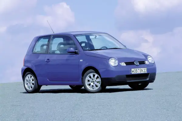 Fioletowy Volkswagen Lupo z tablicą rejestracyjną WOB SN 23 stoi na szarej nawierzchni pod błękitnym niebem z chmurami.