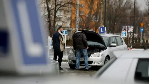 Dwóch mężczyzn przy samochodzie z otwartą maską, znaki "L" i numery na placu manewrowym.