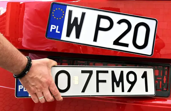Dłoń z bransoletką trzyma tablicę rejestracyjną "O 7FM91". Nad nią druga tablica "W P20". Co oznacza czerwona rejestracja? To pytanie bez odpowiedzi na tym zdjęciu.