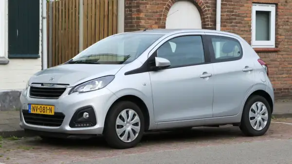 Srebrny Peugeot 108 stoi zaparkowany na brukowanym podjeździe, obok ceglanego domu.