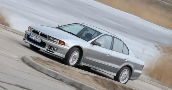Srebrny Mitsubishi Galant z kierowcą za kierownicą, pokonujący zakręt na drodze.