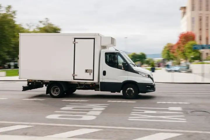 Biała chłodnia na podwoziu Iveco Daily, idealna do transportu wymagającego odpowiedniego prawa jazdy C1, jedzie po miejskiej ulicy.